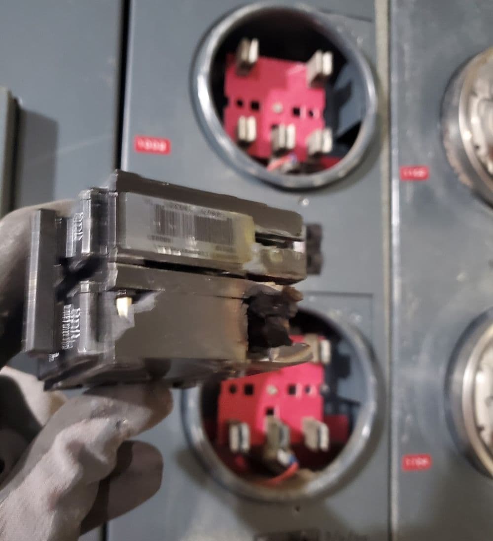 Damaged electrical component held by gloved hand near circuit breakers in power panel.