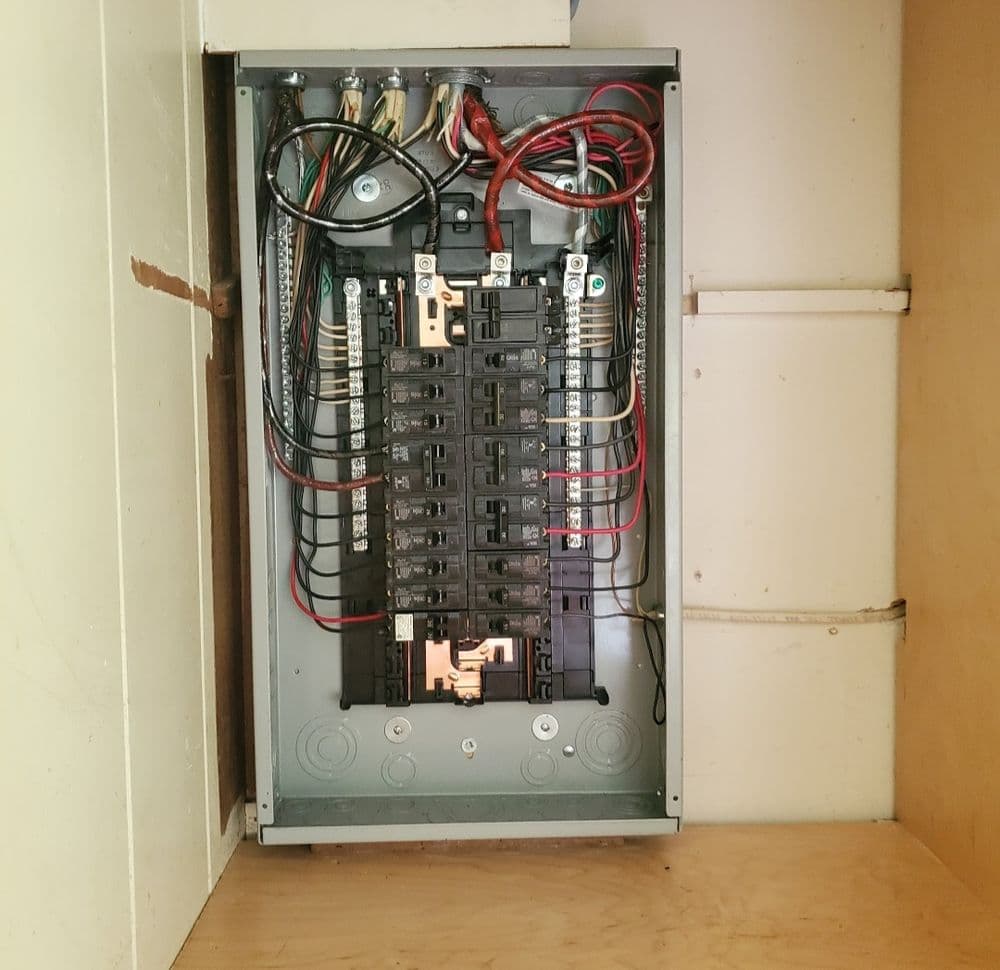 Electrical panel with circuit breakers and wiring, installed in a wooden cabinet.