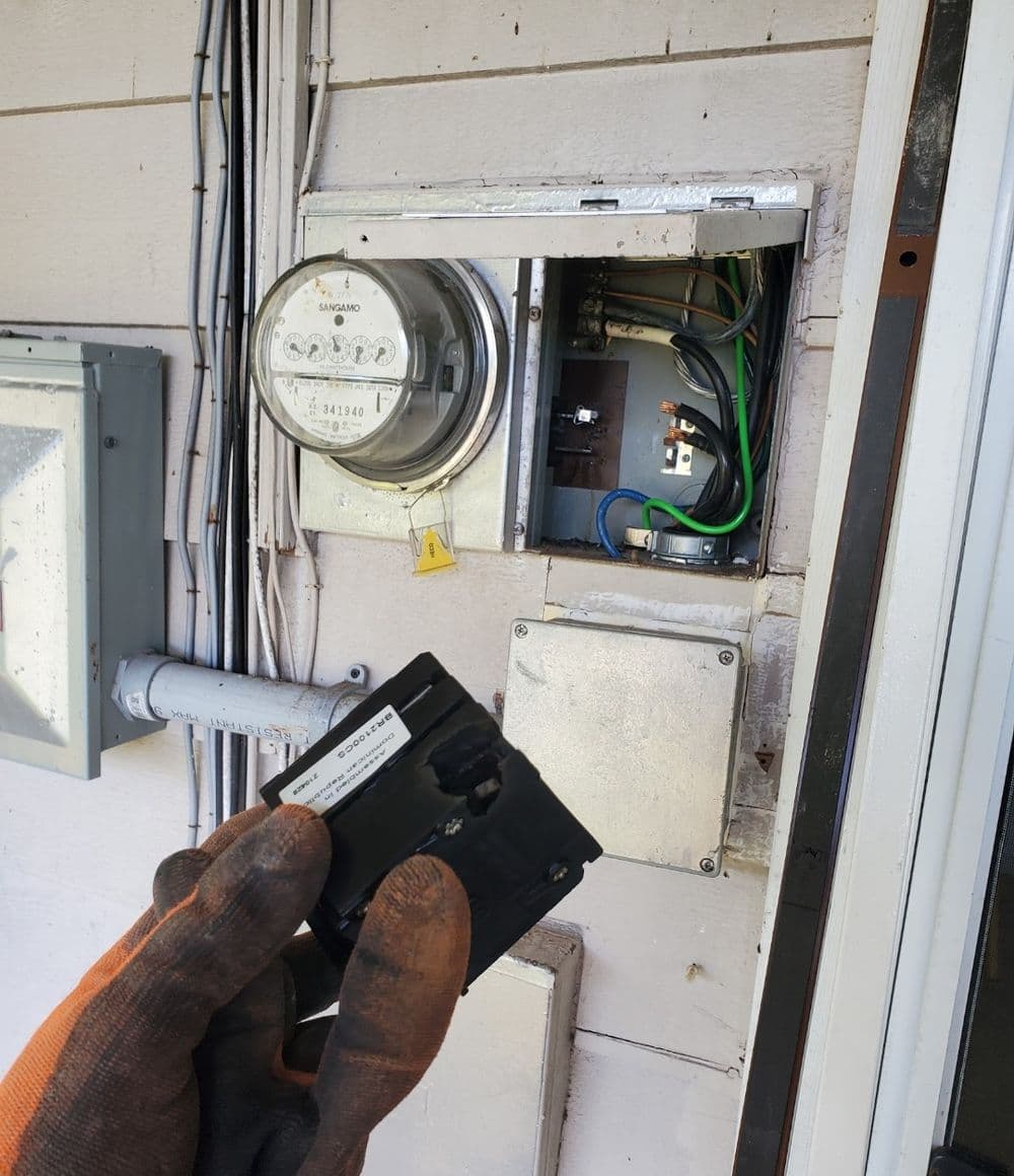 Person holding a device near an electric meter and circuit box with visible wires.