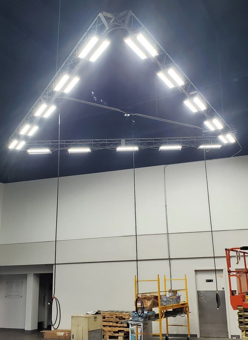 Triangular light fixture hanging from ceiling in industrial space with scaffolding below.
