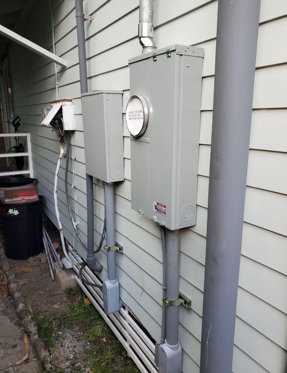 Electrical meter and service panel mounted on gray house siding with conduits and wiring.