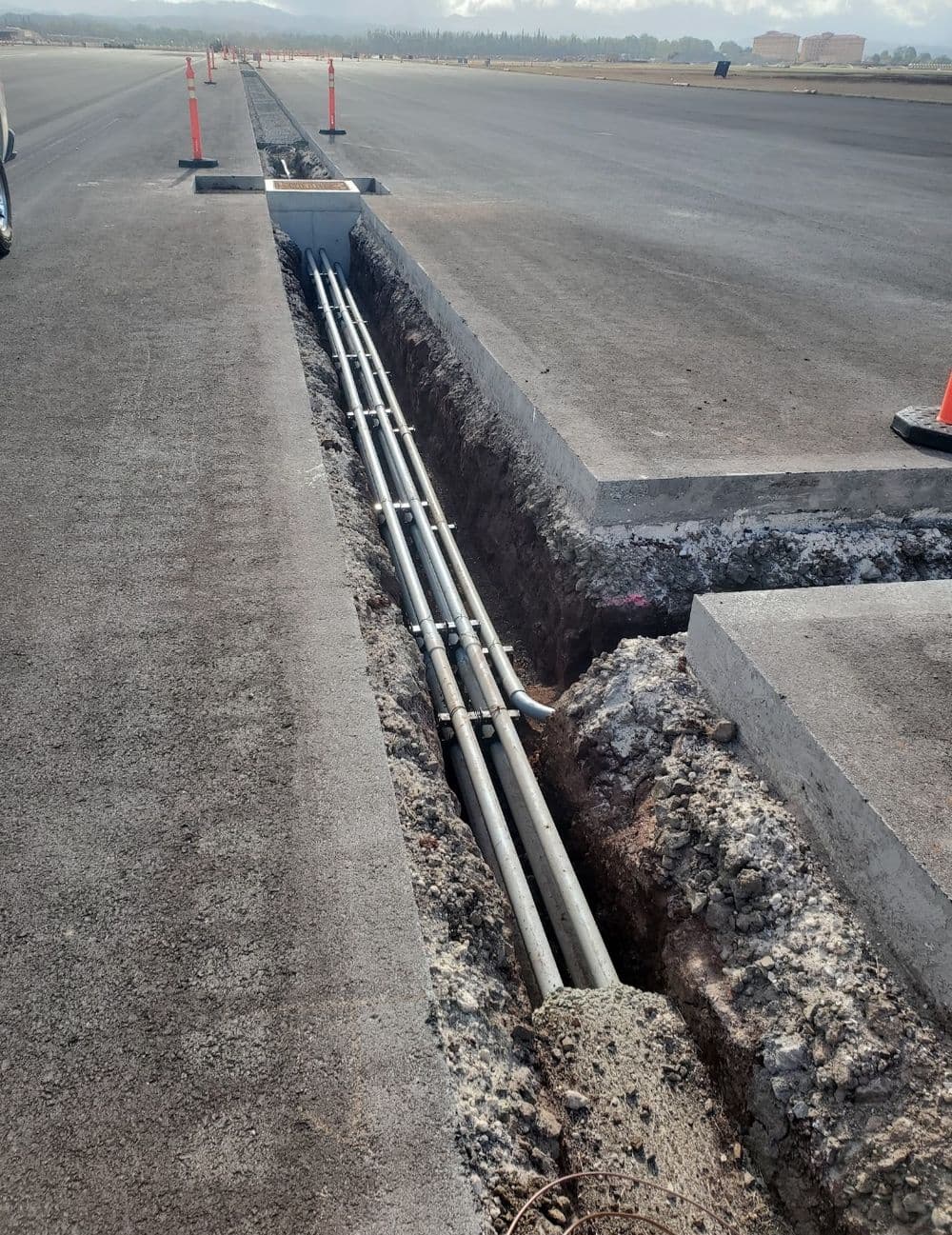 Exposed utility trench with pipes alongside a newly paved road under construction.