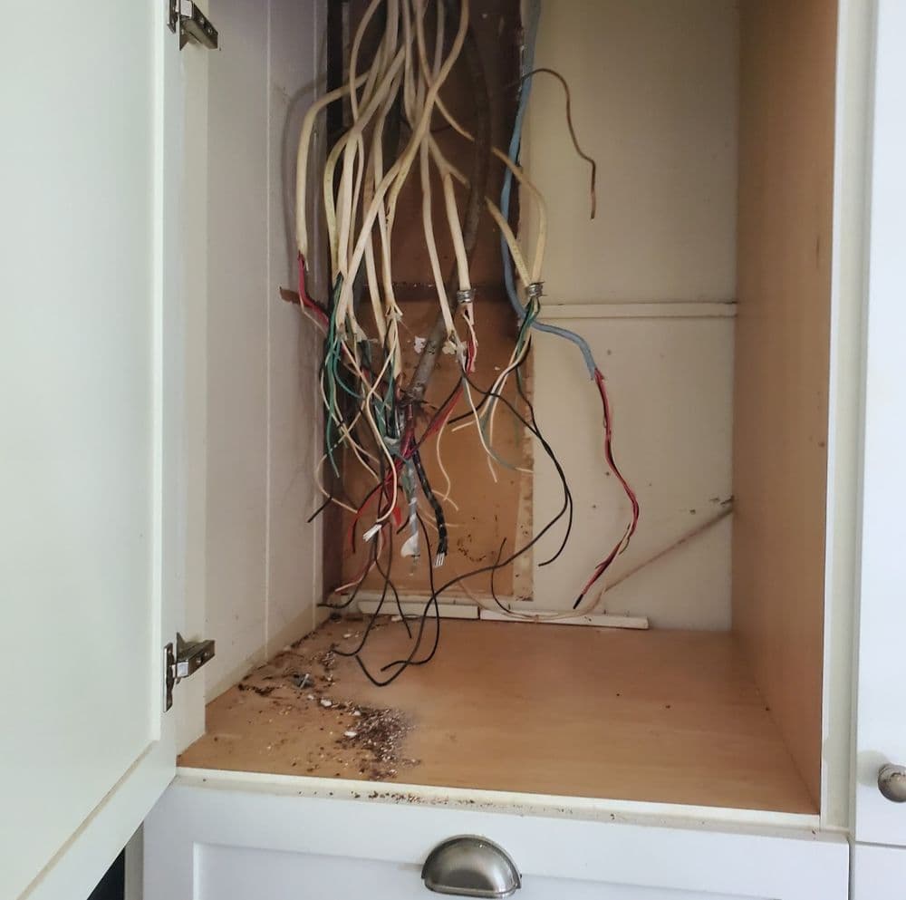 Exposed electrical wires inside a kitchen cabinet with signs of damage and debris.