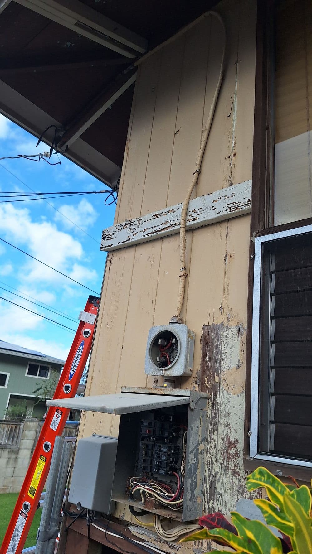 Weathered exterior of a house showing electrical panel, wiring, and a ladder nearby.