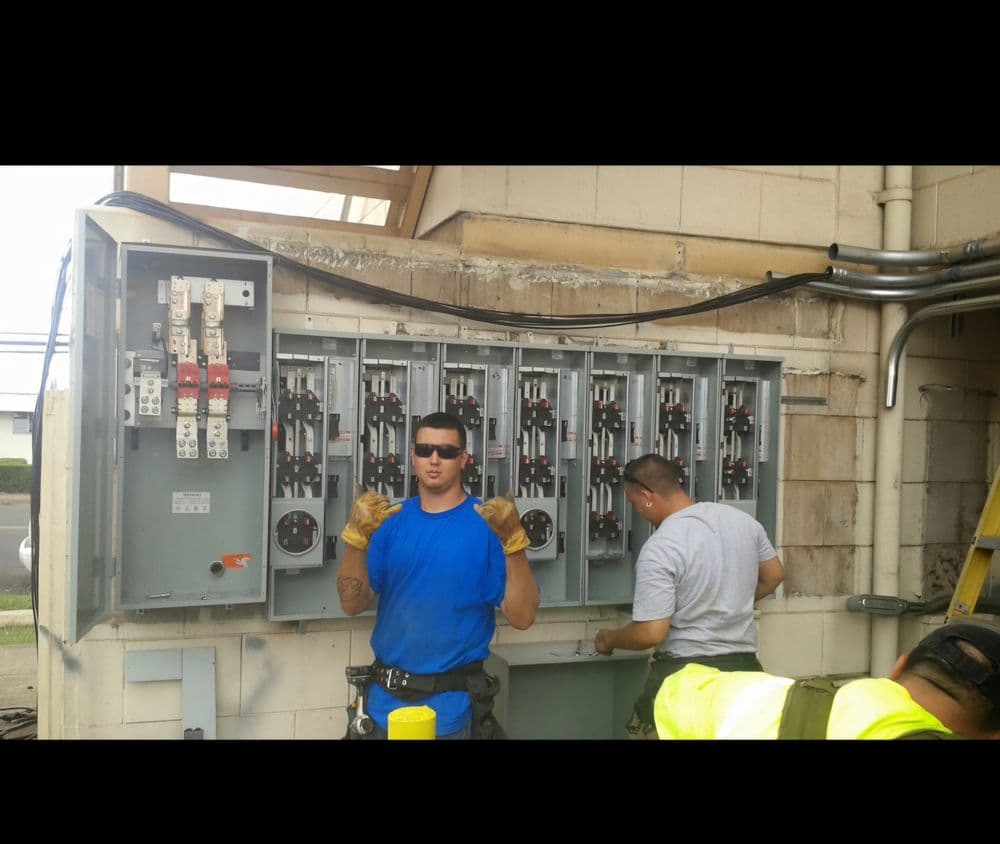 Two electricians working on a large electrical panel outdoors, wearing safety gear.