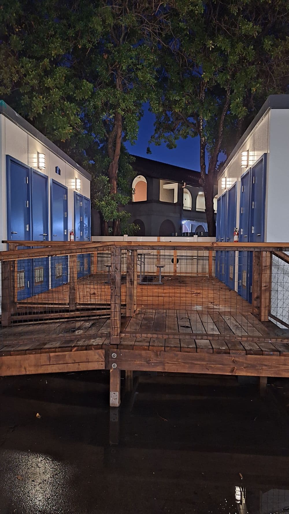 Nighttime view of wooden deck leading to blue storage lockers and surrounding trees.