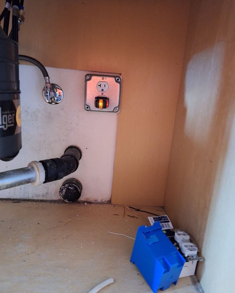 Electrical outlet under sink with plumbing pipes and a blue electrical box.