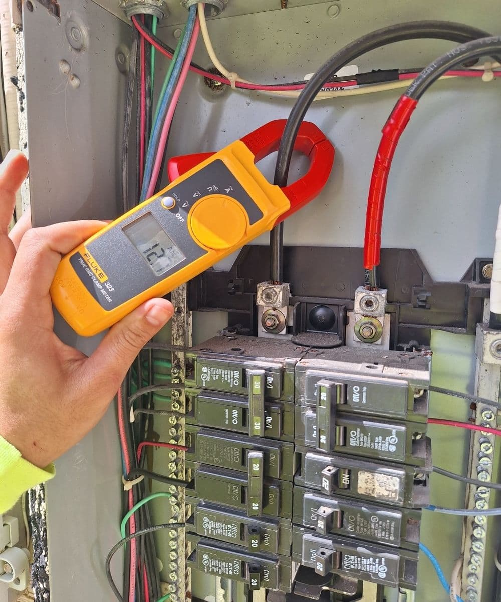 Technician measuring electrical current with a clamp meter on a circuit breaker panel.