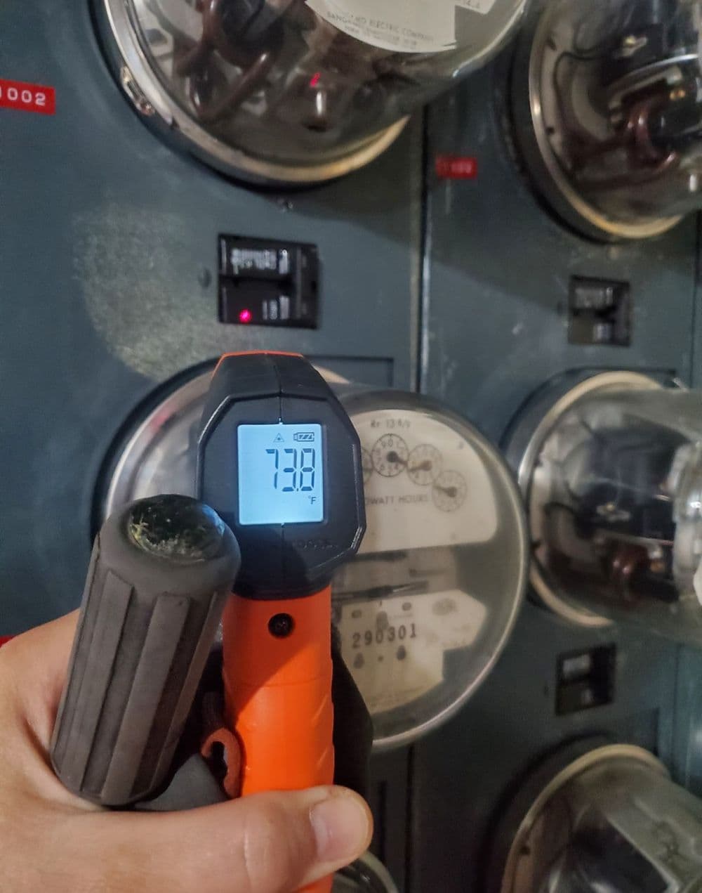 Person holding a digital thermometer reading 73.8°F in front of electric meters.
