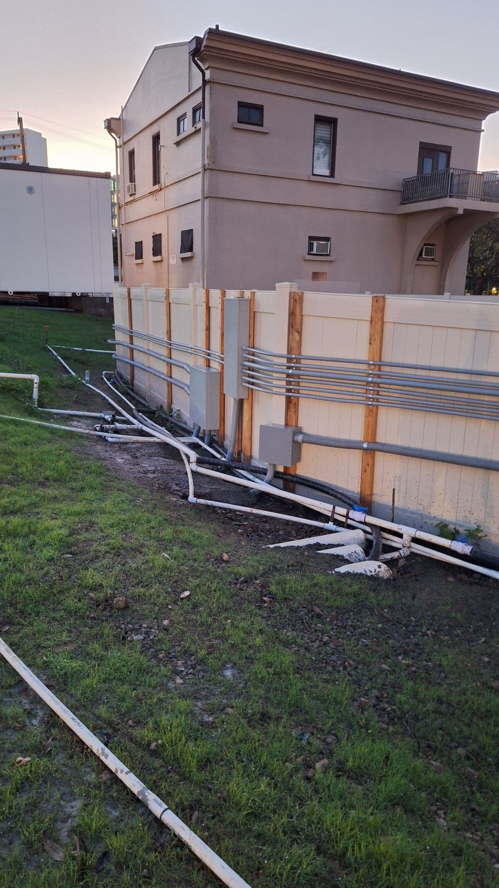 Pipes and electrical lines along a building's fence in a grassy area at sunset.