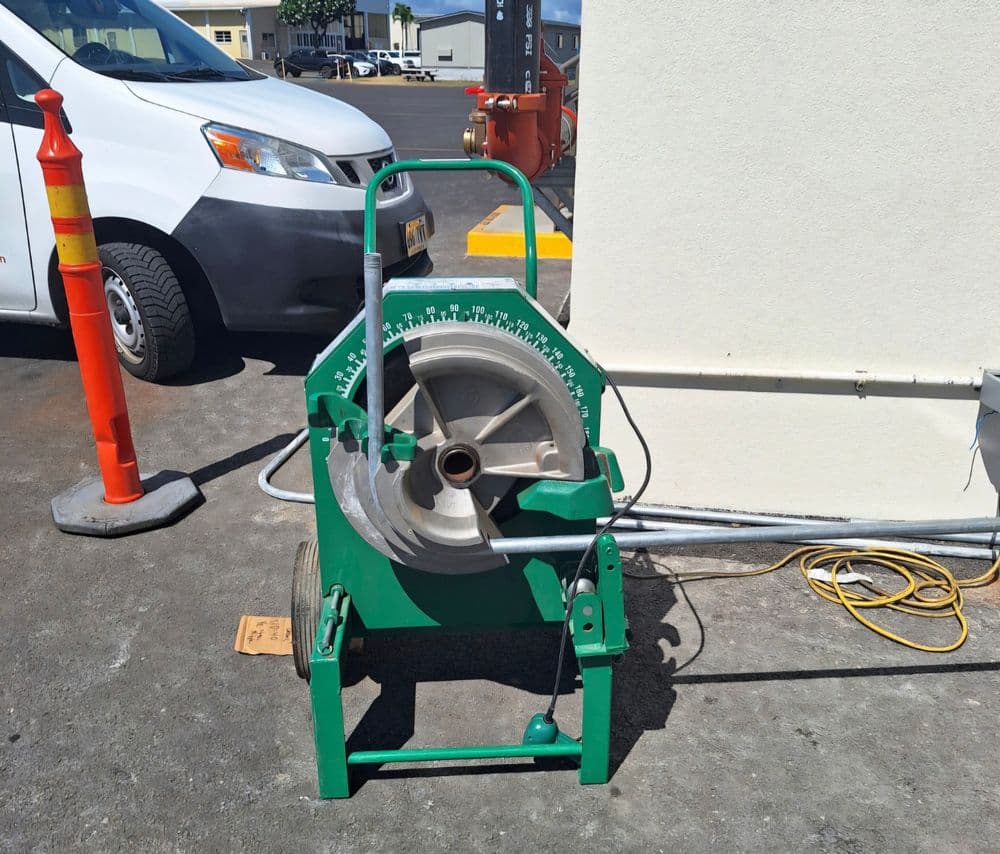Green pipe cutter machine on a parking lot with a vehicle and safety cone in the background.